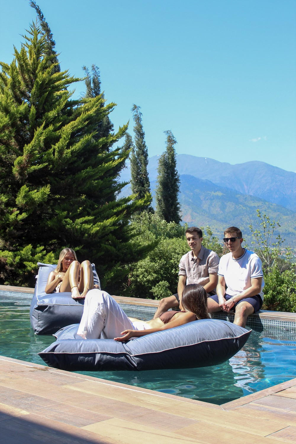 Cuatro personas compartiendo junto a una piscina, dos de ellas sobre cojines Köff flotando, con la cordillera al fondo