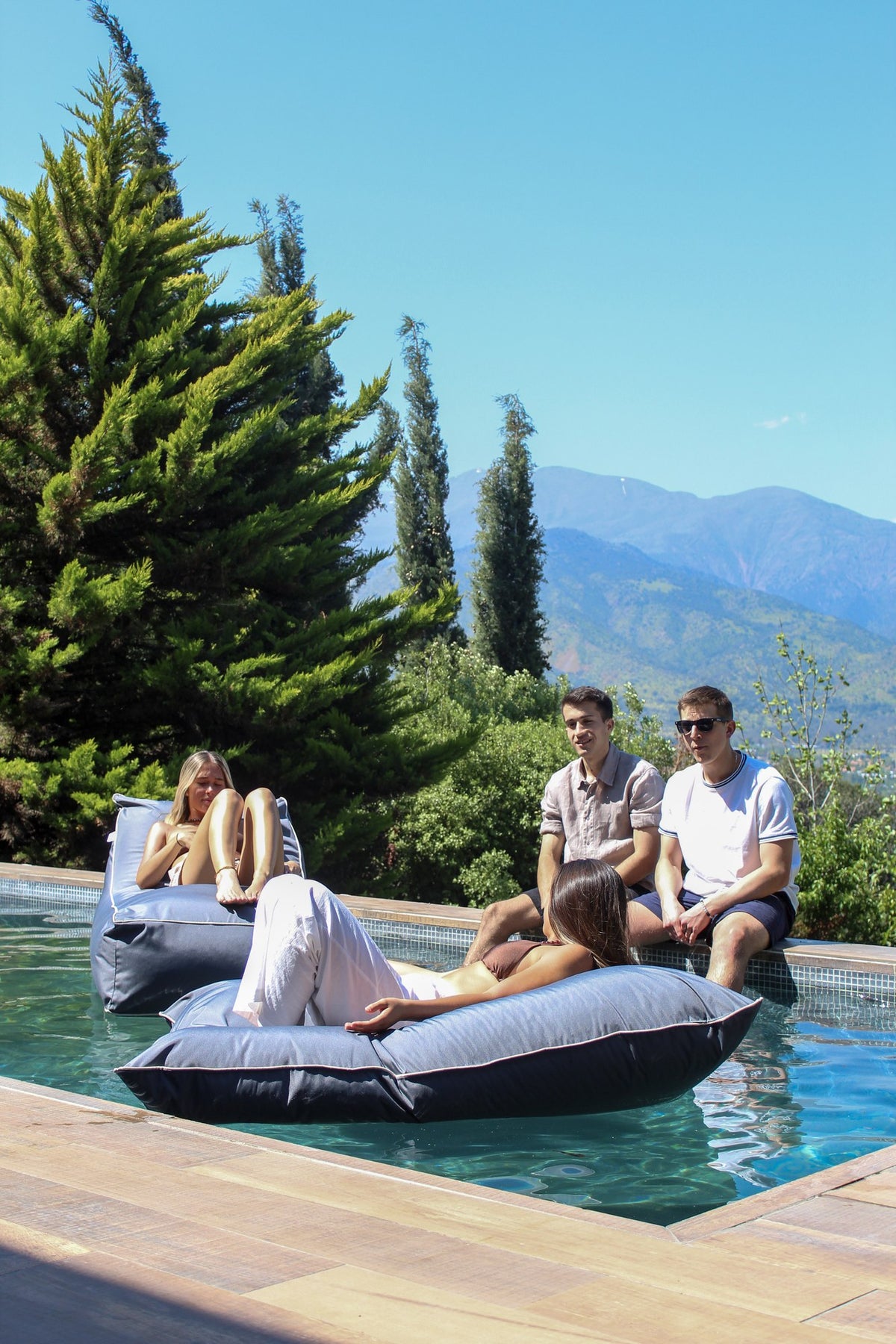 Cuatro personas compartiendo junto a una piscina, dos de ellas sobre cojines Köff flotando, con la cordillera al fondo